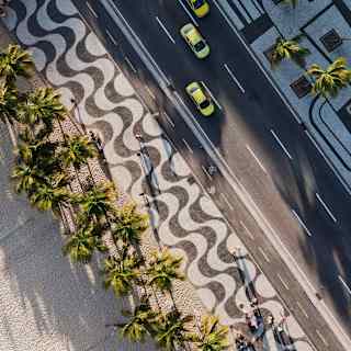 An aerial view shows the iconic black and white wavy lines of the Copacabana boardwalk, flanked by yellow cabs and palms