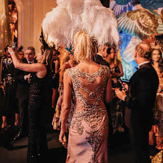 A blonde woman with a lilac lace dress and flamboyant white ostrich-feather headdress, seen from behind at a carnival party.