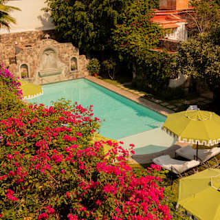 High-angle view over gardens, capturing red blooms, the serene swimming pool, and San Miguel Allende's pink church beyond.