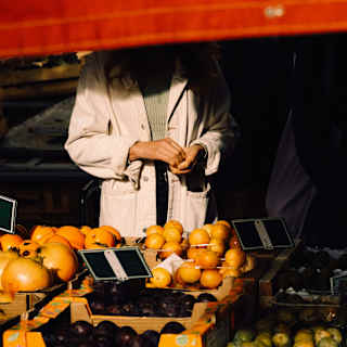 A red awning obscures the face of a man in a white coat who stands at a stall displaying pomegranates, oranges, plums and tomatoes.