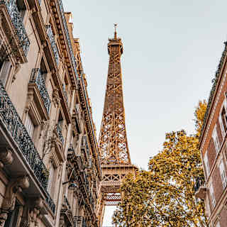Looking up at the Eiffel Tower from a street flanked by tall buildings, one with iron filigree Juliet balcony railings.