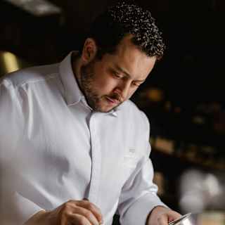 Chef Ricardo Chaneton, with dark hair, short beard and chef whites, carefully spoons sauce from a small pan onto a plate.