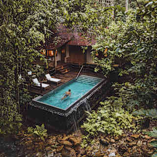 A man swims in a small private pool on a deck surrounded by jungle and overlooking a rock-banked river, seen from above.