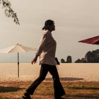A woman in a white shirt and trousers strides across a shadowy grass area behind a beach with a two parasols, red and white.
