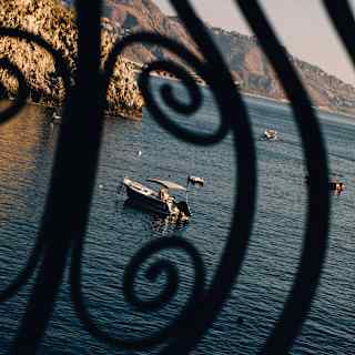 A boat floats on calm sea near a rocky headland, with others further out, glimpsed through curled wrought iron railings.