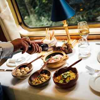 A waiter's hands crack a black pepper mill over four small dishes of brunch food, seen at a window table with a white cloth.