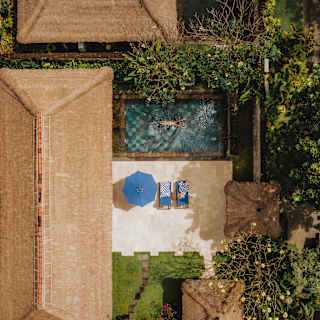 A guest floats in a sapphire pool of a Deluxe Villa, overlooked by a terrace with a parasol and loungers, seen from above.