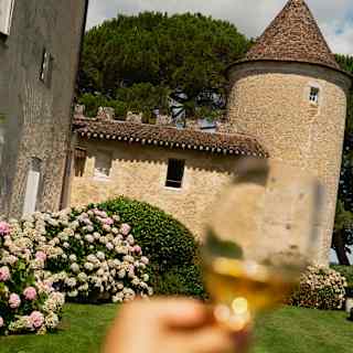 A hand holds a glass of white wine in front of a stone building with a conical turret, surrounded by flowering hydrangeas and green grass.