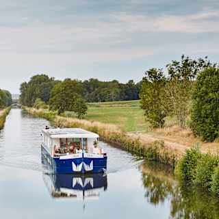A blue and white boat travels down a narrow canal surrounded by green trees and grassy fields under a cloudy sky.