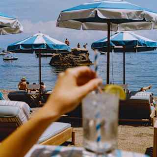A guest stirs a lemon-garnished drink in soft-focus foreground as swimmers gather on a rock out to sea, beyond the parasols.