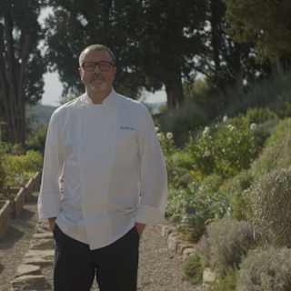 Chef Daniele Sera, with glasses, stands hands-in-pockets in an abundant kitchen garden between raised beds and herb borders.