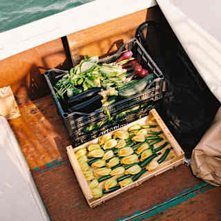A waterborne boat carries two crates in its hull, one with zucchinis and flowers, the other with red onions, seen from above.