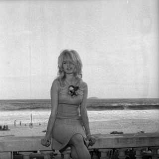 Black and white photo of film star Brigitte Bardot on the wall outside hotel in 1964 in a short dress, with beach backdrop.