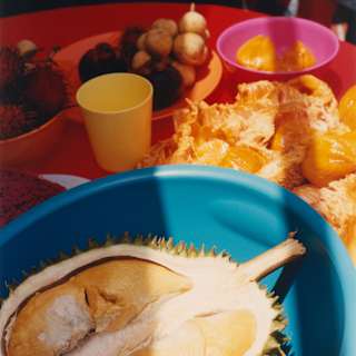 Close-up of a large durian fruit half in a blue bowl on a red table with bright plates of local Malay side dishes.