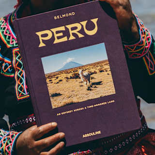 A Peruvian woman in bright traditional woven attire holds a copy of Peru, with a cover-photo of an alpaca, seen close-up.