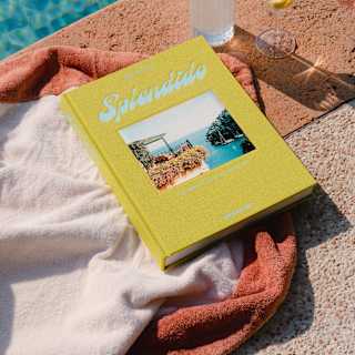 A copy of Splendido, with yellow binding and cover-photo of a bay view rests on a towel with two drinks by a glittering pool.