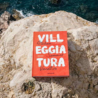 A red book with the title Villeggiatura broken into three lines, and 'Italian Summer Vacation', lies on a rock above the sea.