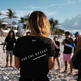 A ring of people wearing Parley For The Oceans tops gather on the beach, seen close-up from behind a blonde woman.