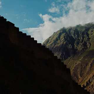 The steps of the Macha Picchu ruins, silhouetted in profile against the green-topped mountains and blue, cloudy sky, behind.