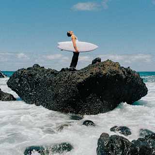 A man with a shortie surfboard under his arm stands, looking at the sky ,on a large basaltic rock surrounded by foaming sea.