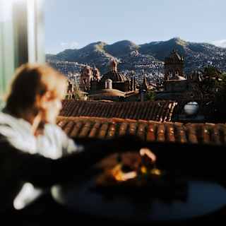A guest in soft-focus foreground gazes through a window at Cusco's terracotta roofs and incredible mountain ranges beyond.