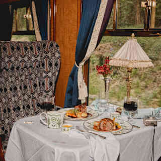 Roast dinner for two rests on a white-clothed window table with red wine and flowers, and a napkin spilling over the edge.