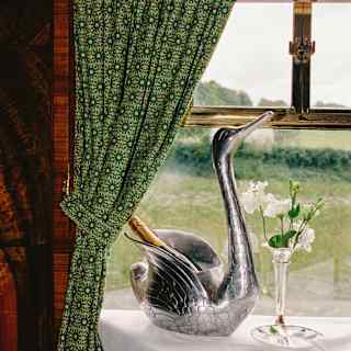 A swan-shaped silver-tone wine cooler holds a bottle of Champagne between its wings, on a window table with green curtains.
