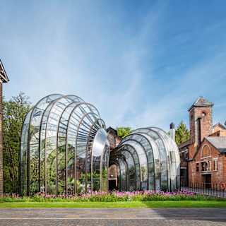 Two intertwined botanical glasshouses with a bulbous funnel design, set on lawned gardens between red-brick buildings.