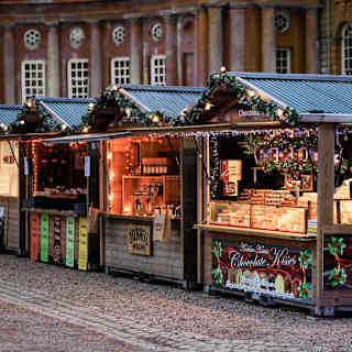 Two women in winter coats browse the pretty illuminated sheds of an artisan Christmas market at Blenheim Palace.