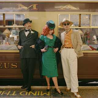 A woman with red curly hair in a green dress and hat leans against the stationed train flanked by two men, all in costume.