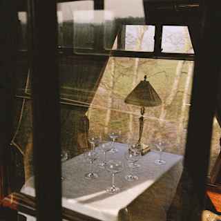 View through a train window into a carriage where eight empty coupe glasses rest on a white-clothed table with a lamp.