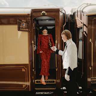 A male steward helps a female guest, wearing a luxurious red dress, step down from the train carriage on to a welcome carpet.