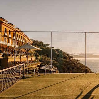 A woman, half-silhouetted against the blaze of the sun, plays tennis on a court overlooking the sea and the hotel and gardens.