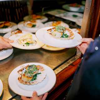 A waiter lifts two plates from the pass, both containing two juicy pan-fried scallops with herb crust, served in the shell.