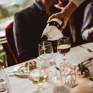 Tilted image of a dining table with water and wineglasses and a side plate of bread, as a waiter pours wine for a male guest.