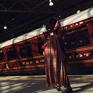 Half-hidden in shadow, a bagpipe player in traditional kilt, plays on a platform in front of the stationary train.