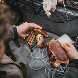 A man holds two large crabs, one in each hand, as he stands by fishing nets and ropes, seen from above, over his shoulder.