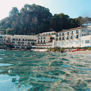 Eye-level view over crystal sea, looking towards Villa Sant'Andrea's white waterfront edifice at the foot of a forested hill.