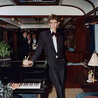 A man in a dinner jacket stands beside a grand piano in a luxurious, wood-panelled train carriage, holding a drink. A person plays the piano whilst others are visible in the background.
