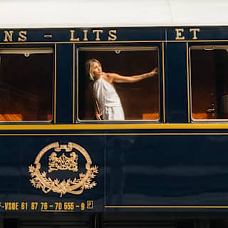 A blond woman in a white halter-neck dress stretches an arm across the train window as she peers out, seen from the platform.