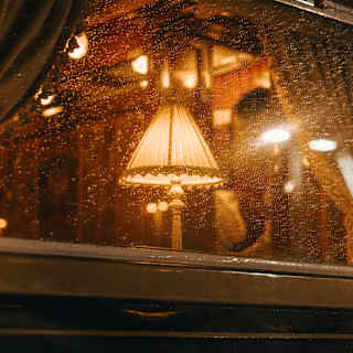 A vintage style lamp glows warm behind a rain-spattered window, seen in an exterior image of the train at night, in close-up.