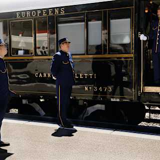 Three male stewards stand on the station platform, with another in a carriage doorway wearing blue uniforms with gold piping.