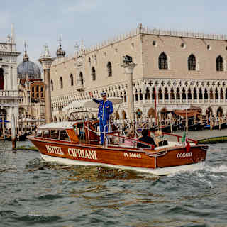 A man in a blue uniform waves as he stands on Hotel Cipriani's wooden boat with three guests, docking by the Doge's Palace.