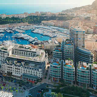 Aerial view of a marina filled with yachts and boats, surrounded by modern and historic buildings, with the sea and hills in the background.