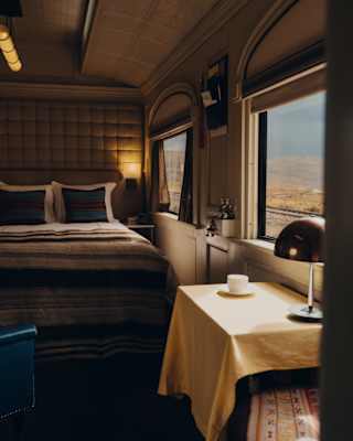 View through a cabin over a window table with a yellow cloth to a double bed with warm, brown woven throws and cushions.