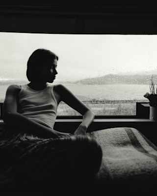 A woman in a vest top leans back against a window that is spattered with rain in a subdued black and white image.