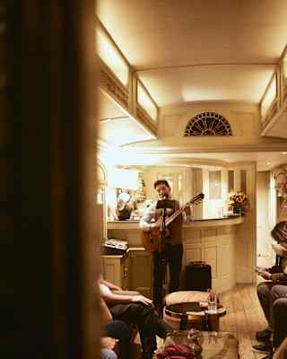 A guitarist stands by the bar, performing for a group, who sit either of a table with drinks, viewed through a car doorway.