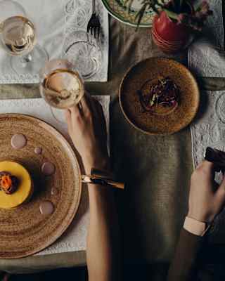 A woman reaches for a glass of wine next to her vibrant dish, in a direct view from above that captures her companion's arm.