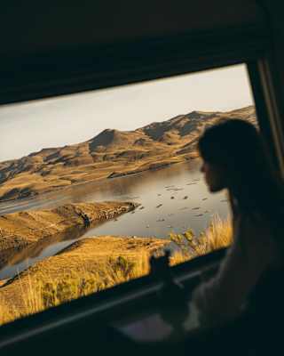Angled image of a woman, in soft-focus, silhouetted against a window with crisp views across a lake edged with gold grasses.
