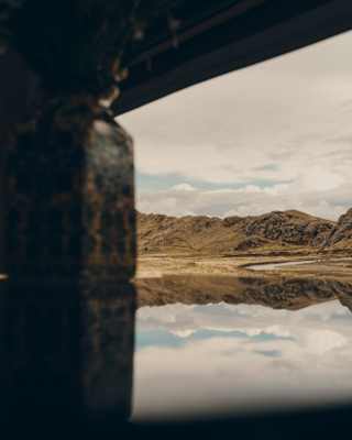 A lake mirrors sky and clouds in a view through a carriage window that captures the rugged, olive green hills behind.